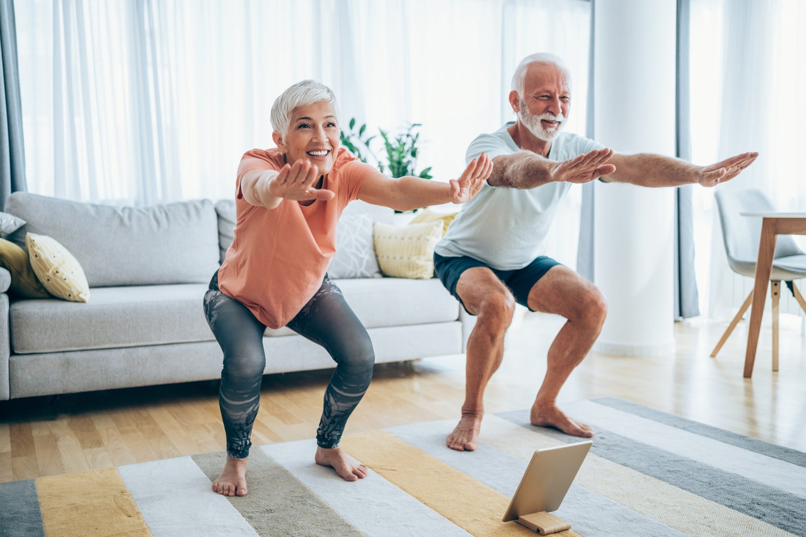 Beautiful mature couple doing workout at home. Couple doing exercises while watching workout video. Senior man and senior woman in sports wear doing workout at home. Couple using digital tablet while doing stretching exercises. Senior couple staying in front of tablet and watching online sport lessons. Couple exercising and watching online fitness live streaming classes.